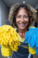 Cheerful mature woman with curly hair holding colorful mops while working as cleaning service professional in office hallway.