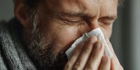 Middle-aged Caucasian man with beard blowing nose into tissue, showing signs of illness, wearing gray scarf, in natural window light.