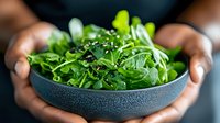 Fresh arugula leaves with sesame seeds served in dark ceramic bowl held by hands. Green salad with peppery taste for healthy eating and cooking.