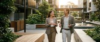 Two professionals walking and conversing in a modern urban courtyard with wooden decking, green plants, and contemporary architecture during golden hour lighting.