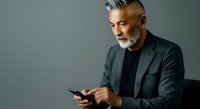 Mature man with gray hair and beard, wearing dark blazer and black shirt, holding smartphone while looking at screen against neutral gray background.