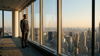 Professional businessman in suit standing by floor-to-ceiling windows overlooking urban skyline at sunset, corporate office setting with city view.