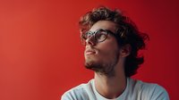 Young Caucasian man with curly hair and glasses wearing white t-shirt looking up thoughtfully against vibrant red background. Professional studio portrait capturing contemplative expression.