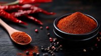 Ground red chili powder in black ceramic bowl and wooden spoon with dried red chilies and peppercorns on dark wooden surface. Food seasoning concept.