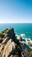Dramatic rocky cliff overlooks turquoise ocean waters with crashing waves below under clear blue sky. Coastal landscape for travel and nature projects.