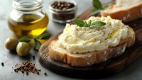 Creamy ricotta cheese spread on rustic bread slice with fresh herbs, olive oil and peppercorns on wooden board for gourmet cooking and healthy eating.