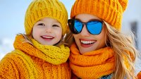Happy Caucasian mother and daughter wearing bright yellow orange knitted winter hats and scarves smiling together outdoors in snow on sunny day.