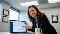 Professional businesswoman pointing at computer screen displaying data analytics dashboard in modern office environment with certificates on wall.