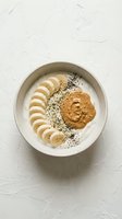 Overhead view of creamy peanut butter yogurt bowl topped with sliced banana, poppy seeds, and granola on white textured surface. Healthy breakfast.