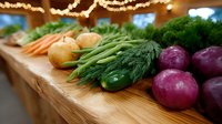 Fresh organic vegetables arranged on rustic wooden table with string lights above, including red onions, cucumbers, dill, carrots and potatoes for farm-to-table dining.