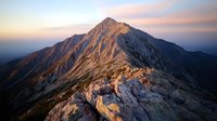 Majestic mountain peak illuminated by warm sunrise light, dramatic rocky ridge leading to summit against pastel sky, rugged alpine landscape in golden hour.