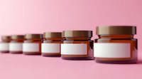 Row of amber glass cosmetic jars with blank white labels on pink background, selective focus. Minimalist packaging mockup for skincare products and beauty branding.