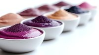 Vibrant natural color pigment powders in white ceramic bowls arranged in row on white background, featuring purple, pink, orange and black mineral dyes for cosmetics.