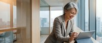 Mature businesswoman with gray hair smiling while using tablet in modern office with city view. Professional corporate executive working on digital device.