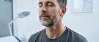 Middle-aged man with gray beard and dark hair wearing casual black t-shirt in modern office or medical setting with blurred background equipment.