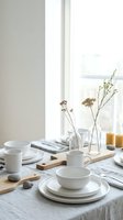 Minimalist table setting with white ceramic dishes, dried flowers in glass vases, and natural linen tablecloth by bright window for dining decor.