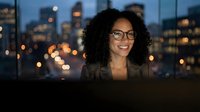 Young woman with curly dark hair and glasses smiling at camera against illuminated city skyline at dusk, suitable for corporate, business, and professional website content.