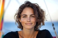 Young Mediterranean woman with short wavy brown hair and natural makeup smiling confidently outdoors against blurred nautical background, radiating joy and authenticity.