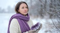 Young Caucasian woman in purple scarf and cream sweater holding snow in winter forest. Peaceful snowy day outdoor portrait for seasonal lifestyle content.