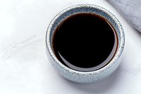 Black soy sauce in traditional blue ceramic bowl on white background, top view close-up showing liquid surface reflection and textured pottery glaze.