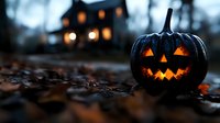 Glowing carved pumpkin with scary face on dark autumn ground with fallen leaves and spooky house silhouette in blurred background with warm lights.