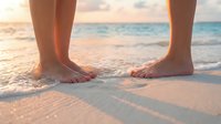 Bare feet standing on sandy beach at sunset with gentle waves washing over toes creating peaceful vacation moment for wellness and relaxation concepts.