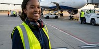 Young African American female airport ground crew worker smiling at camera wearing safety vest and headphones on busy tarmac with aircraft background.