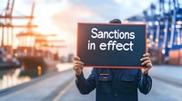 Person holding black sign with text sanctions in effect at industrial port during sunset, cargo containers and cranes in blurred background, trade restrictions concept.