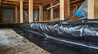 Black vapor barrier plastic sheeting installed on dirt crawl space floor with wooden support beams and joists visible in residential construction project.