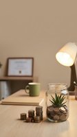 Cozy home office workspace with glass jar savings bank containing coins and small plant on wooden desk with warm lamp lighting and certificate.