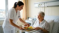 Young female nurse in white uniform serving healthy meal to elderly Caucasian woman patient in hospital bed, healthcare nutrition and medical care service.