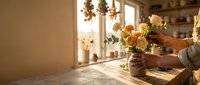 Woman arranging fresh yellow roses in wicker basket on wooden table in sunlit kitchen interior for home decoration and floral design projects.