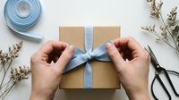 Hands tying blue ribbon bow on kraft paper gift box surrounded by dried flowers and scissors on white surface for present wrapping and holiday gifting.