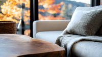 Cozy autumn interior with beige sofa, gray throw pillow and wooden coffee table near window with fall foliage view, creating warm home atmosphere.
