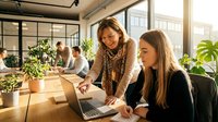 Two young women collaborating at laptop in bright modern office with plants and colleagues working in background, suitable for business, teamwork, startup, and corporate training materials.