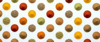 Colorful spice powder circles arranged in repeating pattern on white background. Various cooking seasonings including turmeric, paprika, cumin creating vibrant culinary design.