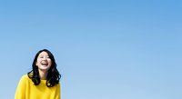 Young woman with long dark hair and East Asian ethnicity wearing a bright yellow shirt, smiling joyfully against a clear blue sky background for lifestyle and happiness concepts.