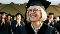 Senior woman graduate smiling proudly in cap and gown at university commencement ceremony with classmates applauding in background for academic achievement.