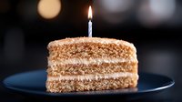 Slice of layered vanilla cake with cream filling and one burning birthday candle on dark blue plate against blurred bokeh background. Celebration moment.