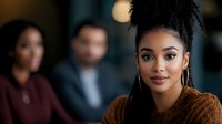 Young Black woman with braided hair styled in a top knot, wearing gold hoop earrings and a warm brown sweater, smiling at camera in modern office setting with blurred colleagues in background.