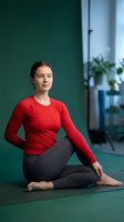 Young Caucasian woman in red long sleeve top practicing yoga meditation pose on mat in modern fitness studio with green wall and plants in background.