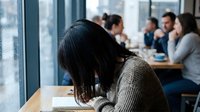 Young Asian woman studying alone at cafe table while diverse group socializes in background, depicting isolation and focus in busy environment.