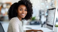Young African American businesswoman with curly hair smiling at camera while working on computer with digital interface icons floating in modern office space.