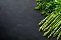 Fresh green asparagus spears and parsley leaves arranged on dark slate background with copy space, raw organic vegetables for healthy cooking.