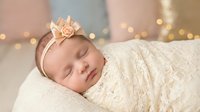 Peaceful newborn baby girl sleeping wrapped in cream lace blanket wearing floral headband with soft golden bokeh lights background for nursery decor.