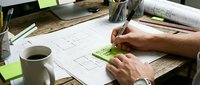 Architect or designer with dark skin tone working on blueprint sketches at wooden desk with coffee cup, drafting tools, and construction plans for architectural design projects.