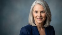 Mature woman with long gray hair and fair complexion wearing dark blue blazer, smiling at camera against neutral gray background for corporate portraits and professional headshots.