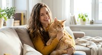 Young woman with long brown hair cuddling orange tabby cat on white sofa in bright modern living room with plants and natural light streaming through windows.