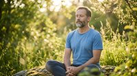 Middle-aged Caucasian man in casual blue t-shirt meditating outdoors in natural green garden setting during golden hour sunlight for wellness and mindfulness content.