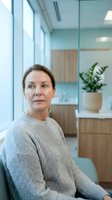 Middle-aged Caucasian woman in gray sweater sitting in modern office space with glass walls and natural light for business and professional concepts.
